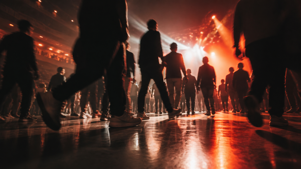 Anonymous crowd silhouettes cross an arena floor under orange lights.